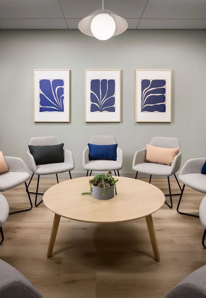 Group room with grey armchairs in a circle around a round wooden table, under three blue botanical prints